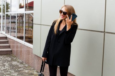A slender young smiling blonde in stylish clothes, a black jacket and leggings, in sunglasses, with a leather bag in her hands, stands on the street near a modern building and talks on the phone.