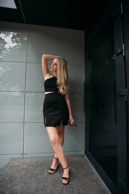 A stylish slender girl with blond long hair and long legs stands against the backdrop of a modern building. Dressed in a short black sexy skirt and top.