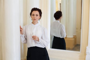 A portrait of a respectable woman in ancient Ukrainian clothes, in a black long skirt and a white blouse, stands in the premises of a chic baroque estate, with large columns and a mirror.