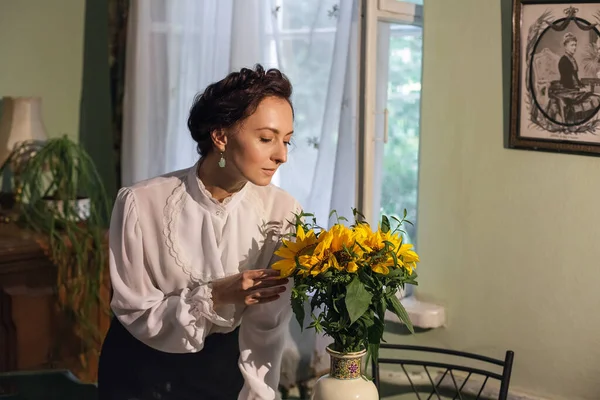 A stylish Ukrainian woman in vintage clothes, a black skirt and a white shirt, stands by the window in a cozy apartment with retro furniture. There is a vase with sunflowers on the table.