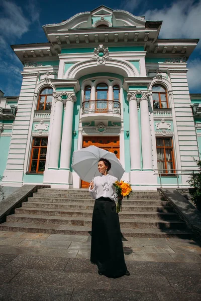A stylish Ukrainian woman in vintage clothes, a black skirt and a white shirt, stands with an umbrella, near the facade of an old estate with beautiful architecture, on a bright sunny day.