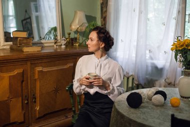 A stylish Ukrainian woman in vintage clothes sits at a table in a cozy apartment with retro furniture, holds coffee in her hands. There is a vase with sunflowers on the table.