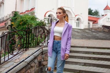 A beautiful girl with blond hair, in a purple jacket and a white top and jeans, with chic makeup and a stylish hairstyle, stands on the stairs, against the backdrop of beautiful architecture.
