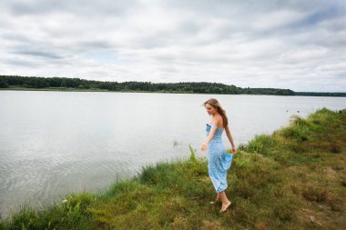 A beautiful slender smiling girl with long blond hair, in a blue summer dress, spins and walks along the river bank, against the backdrop of a picturesque landscape.