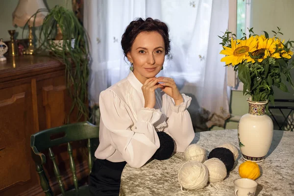 A stylish Ukrainian woman in vintage clothes sits at a table in a cozy apartment with retro furniture. There is a vase with sunflowers and balls of thread on the table.
