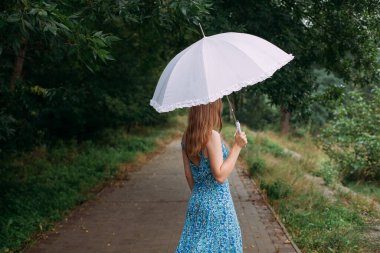 A slender young girl with blond long hair, in a blue summer dress, stands with a white umbrella in her hands, on a path in a forest park area in the rain.