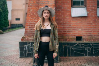 A slender young girl with blond long hair, in black leggings, a tank top, a beige jacket and a hat, stands on the street near the wall of an old building.
