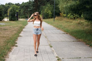 Uzun sarı saçlı, kısa seksi kot şortlu, beyaz bluzlu, güneş gözlüklü, parkta yürüyen ince bir kız..