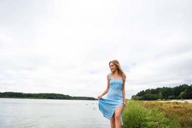 A beautiful slender girl with long blond hair, in a blue summer dress, walks on the water, in the river, against the backdrop of a picturesque landscape.