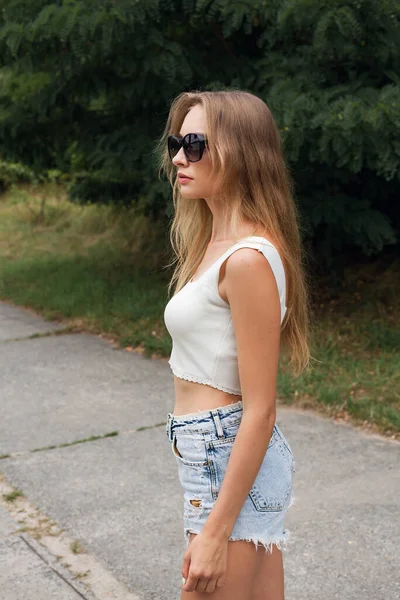 A young slender girl with long blond hair in short sexy denim shorts and a white top, in sunglasses, stands on a path in the park.