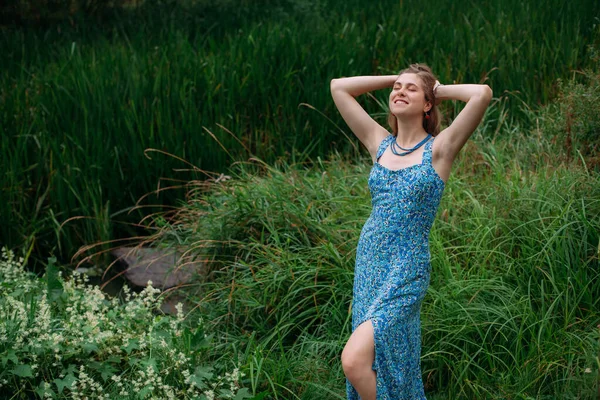 A slender young girl with blond hair, in a blue summer dress, stands on a green lawn in a forested area and smiles.