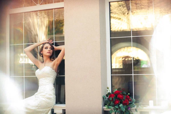 レストランの窓に座っている明るいウェディングドレスを着たスタイリッシュな美しい花嫁の肖像画であるアート作品は 花のシックなインテリアを背景にしています — ストック写真