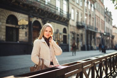 Güzel, şık giyinmiş, sarı saçlı bir kız güneşli bir günde eski şehrin sokaklarında, güzel binaların etrafında dikiliyor..