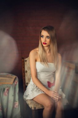 Parlak makyajlı, uzun sarı saçlı, açık renk elbiseli güzel bir kız bir masada oturuyor..