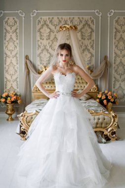 Şık makyajlı, beyaz bir gelinlik giymiş güzel bir gelin, düğün öncesi sabah şık bir iç mimariye sahip bir yatak odasında duruyor..