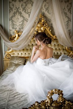 Şık makyajlı, beyaz bir gelinlik giymiş güzel bir gelin, düğün öncesi sabah şık bir iç dekorasyonla bir yatak odasında oturur..