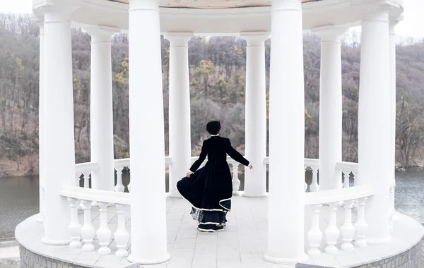 A slender woman in a stylish vintage look, a long black dress and a stylish hairstyle, dances in a white gazebo with columns in winter.