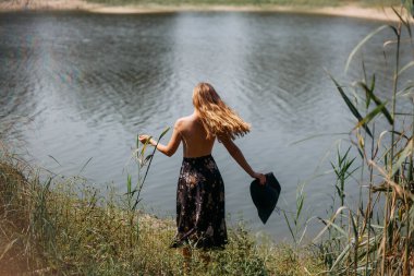Uzun sarı saçlı, uzun etekli ve elinde geniş siyah şapkalı ince bir genç kız, çimlerin üzerinde dönüyor, pitoresk nehrin yanında..