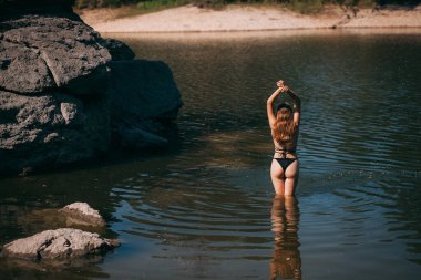 Uzun sarı saçlı, ince vücutlu ve güzel sırtlı, siyah mayo giymiş genç bir kız, kayaların arkasında bir nehirde duruyor..