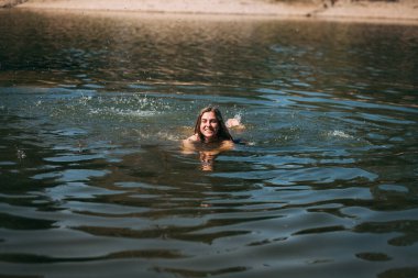 Uzun sarı saçlı genç bir kız, ince bir figür, siyah bir mayo giymiş, yüzüyor ve gülümsüyor, plajın arka planında, sıcak bir yaz gününde..