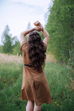 Uzun siyah saçlı bir kız arkası bize dönük, kahverengi bir yaz elbisesi içinde ellerini havaya kaldırıyor, tarlanın ve ormanın etrafında..