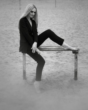 Sanat eserleri. Siyah beyaz bir fotoğraf, güzel bir stil kızı, sarı saçlı, siyah klasik takım elbiseli, plajdaki ahşap bir bankta oturuyor..