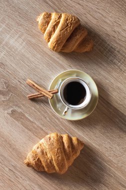 Bir fincan sıcak kokulu siyah kahve ayçöreği ve tarçın çubuklarıyla bir fincan çay tabağında, açık kahverengi ahşap bir masada..