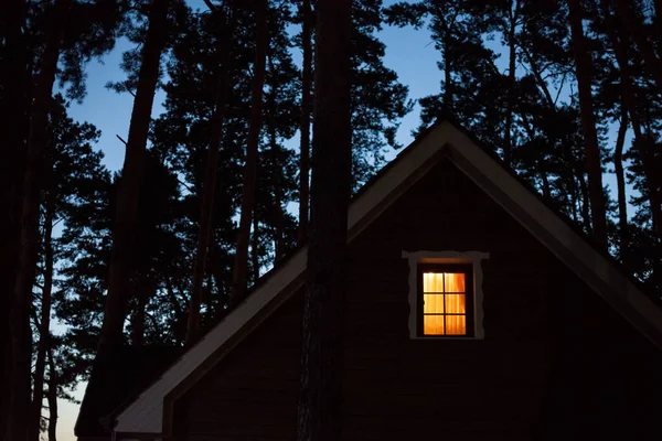 Warm light in the window of a village house at night in the forest at dusk, against the backdrop of trees and a blue sky.