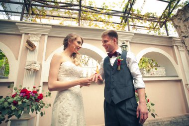 Bir düğün töreninde şık yeni evliler, bir restoranda, yüzükler takıyorlar. Gelin uzun, açık renk bir elbise, damat beyaz bir gömlek ve gri bir kravat giyiyor..