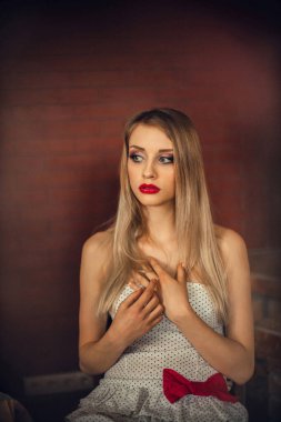 Sarışın, uzun saçlı ve parlak makyajlı zarif genç bir kız, odasındaki bir sandalyede, bordo bir duvarın arkasında oturuyor..