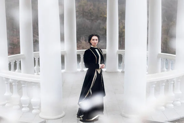 A beautiful woman with a stylish hairstyle and make-up, in a vintage black dress, stands near the columns in a white gazebo in winter. Snow is falling outside.