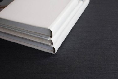 Deri kapaklı, farklı boyutlarda üç adet beyaz renkli fotoğraf kitabı bir odada koyu gri bir yüzeye uzanıyor..