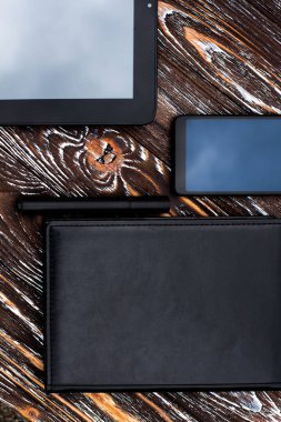 Siyah bir tablet, bir cep telefonu, deri kaplı bir defter ve koyu ahşap bir yüzeye uzanan bir kalem..