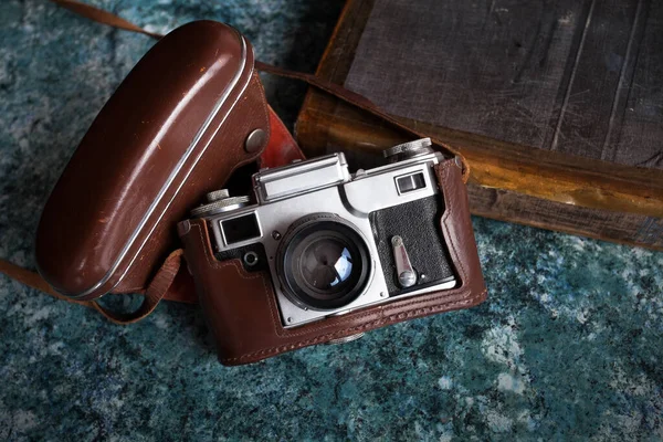Kahverengi deri bir çantanın içindeki eski bir Sovyet kamerası desenli mavi bir masanın üzerinde duruyor. Yakınlarda eski bir kitap var..