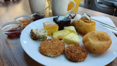 Beyaz porselen tabakta tatlılar. Doğulu kurabiyeler. Hindistan cevizli çikolata küpleri. Cevizli Kadaif. Çörekler. Susamlı kek. Meyve reçelli kavanozlar. Portakal suyu ve kahve. Ahşap arka planda kahve ve kek. Beyaz peçete. Ananas dilimleri..