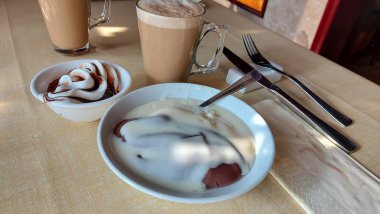 Cam bardakta kahve. Çikolata soslu kremalı dondurma. Masa örtüsü üzerinde bir tatlı. Görüntüyü yaklaştır. Peçetenin yanında çatal bıçak takımı var. Beyaz porselen kase. Restoran sahnesi pencerelerin yanında. Gün ışığı.