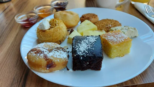 Beyaz porselen tabakta tatlılar. Doğulu kurabiyeler. Hindistan cevizli çikolata küpleri. Cevizli Kadaif. Çörekler. Susamlı kek. Meyve reçelli kavanozlar. Portakal suyu ve kahve..