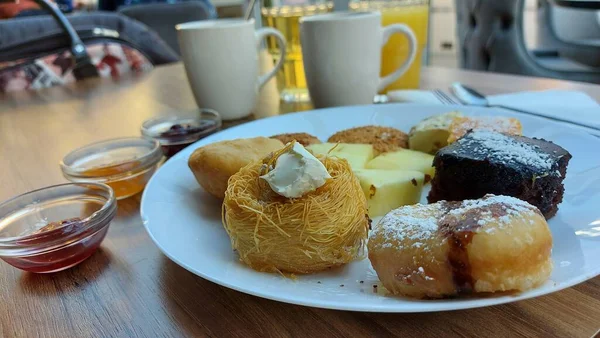 Beyaz porselen tabakta tatlılar. Doğulu kurabiyeler. Hindistan cevizli çikolata küpleri. Cevizli Kadaif. Çörekler. Susamlı kek. Meyve reçelli kavanozlar. Portakal suyu ve kahve..