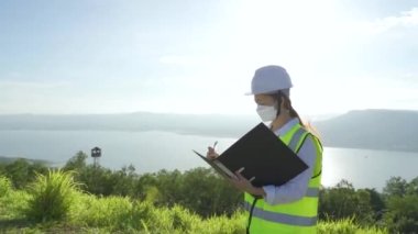 Maskeli ve beyaz kasklı genç mühendis veya teknisyen, yenilenebilir enerji çiftliğindeki belgeyle rüzgar türbini sistemini kontrol ediyor.