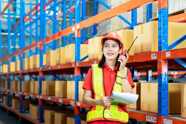 Warehouse logistic management business. young Asian woman wearing reflective safty vest and white helmet, Communicating via walky-talky in the warehouse factory as worker