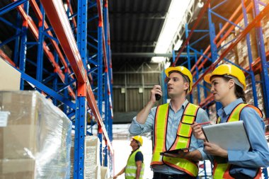 Group of warehouse workers with hardhats and reflective jackets using tablet, walkie talkie radio and cardboard while controlling stock and inventory in retail warehouse logistics, distribution center