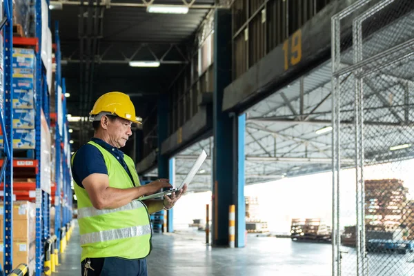 Warehouse Worker Uses Laptop. He\'s Standing in the Middle of a Big Distribution Center with Big Storage Racks and Pallets on Them.