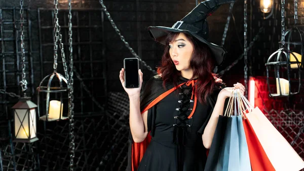 Happy halloween, young asian woman in witch costume show mobile and holding shopping bags. Emotion woo