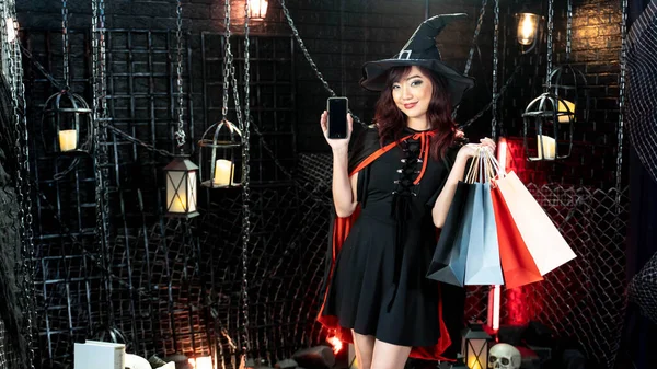 Happy halloween, young asian woman in witch costume show mobile and holding shopping bags.