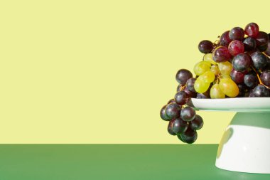 Koyu yeşil arka plana karşı beyaz pasta standı üzerinde taze üzümlerle yapılan yaratıcı meyve düzenlemesi. En düşük sonbahar konsepti.
