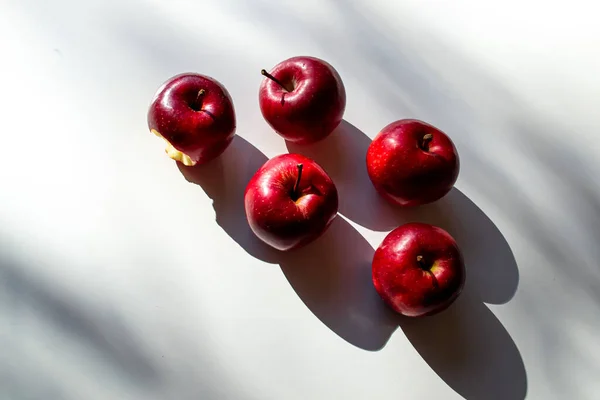 Four whole and one bitten red apples on a white table.