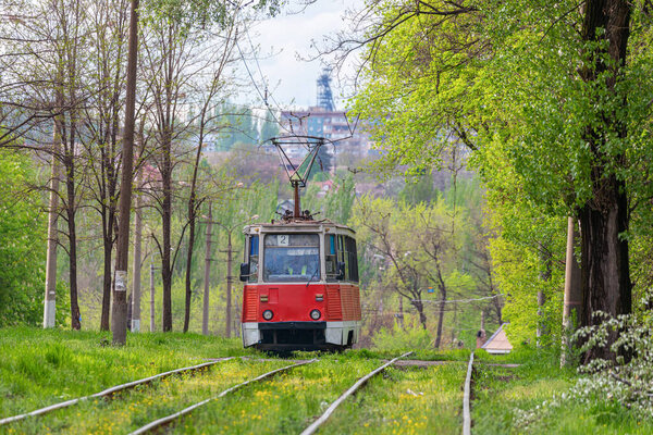An old red tram rides on rails through the city park. Trees with green leaves. Urban passenger transportation.