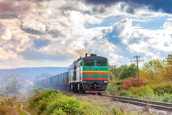 Güçlü yeşil bir dizel lokomotif, kömürlü uzun bir yük trenini tren istasyonuna çeker. Demiryolu taşımacılığı. Bulutlu gökyüzü.