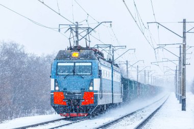 Güçlü gri bir lokomotif tren istasyonuna uzun bir yük treni çeker. Kar fırtınası. Demiryolu taşımacılığı.
