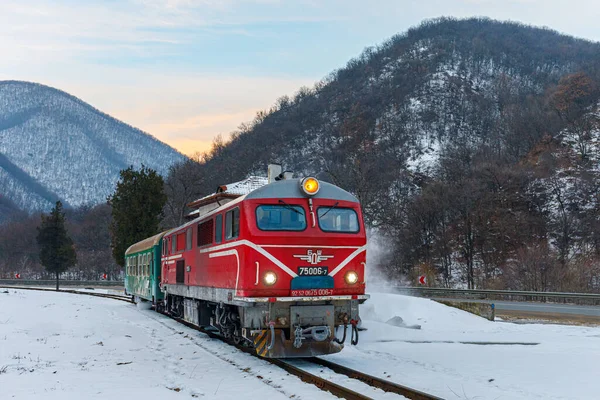 Bulgaristan 'ın Bansko kenti - Kış 2022: Güçlü kırmızı dizel lokomotif bir yolcu trenini raylar boyunca çekiyor. Çok fazla kar var. Arka planda sıradağlar var. Bulutlu bir kış sabahı. Bulgar demiryolu